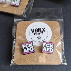 Könnte beinhalten: Quadratische schwarz-weiße Anstecknadeln mit dem Text "FCK AFD" in einem rosa Rand, verpackt auf einer braunen Karte mit dem Text "VGNX PRINTS". Mehrere Pakete sind sichtbar.
