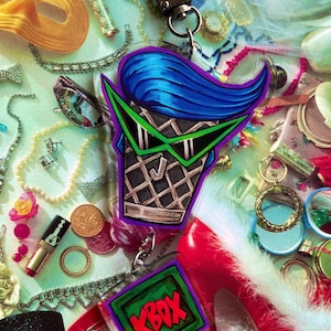 May include: A clear acrylic keychain featuring a stylized ice cream cone with blue hair and green sunglasses. The keychain has a silver-colored metal clasp and a green and red KBOX logo. The background includes jewelry and a red high heel.