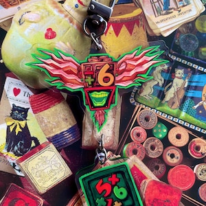 May include: A clear acrylic keychain with a colorful, winged design featuring the number 6 and a green-toothed face. The keychain has a silver clasp and is surrounded by vintage ephemera, including playing cards and wooden spools.