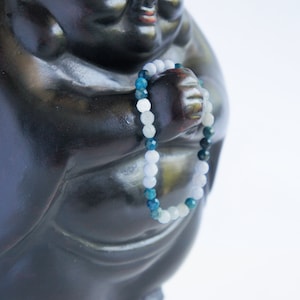 May include: A blue and white beaded bracelet with a dark blue, green, and white color gradient. The bracelet is wrapped around the hand of a dark gray statue.