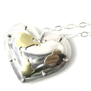 Puede incluir: Un collar con un colgante en forma de coraz&oacute;n plateado con un dise&ntilde;o de mapa dorado. El collar tiene una cadena delicada.