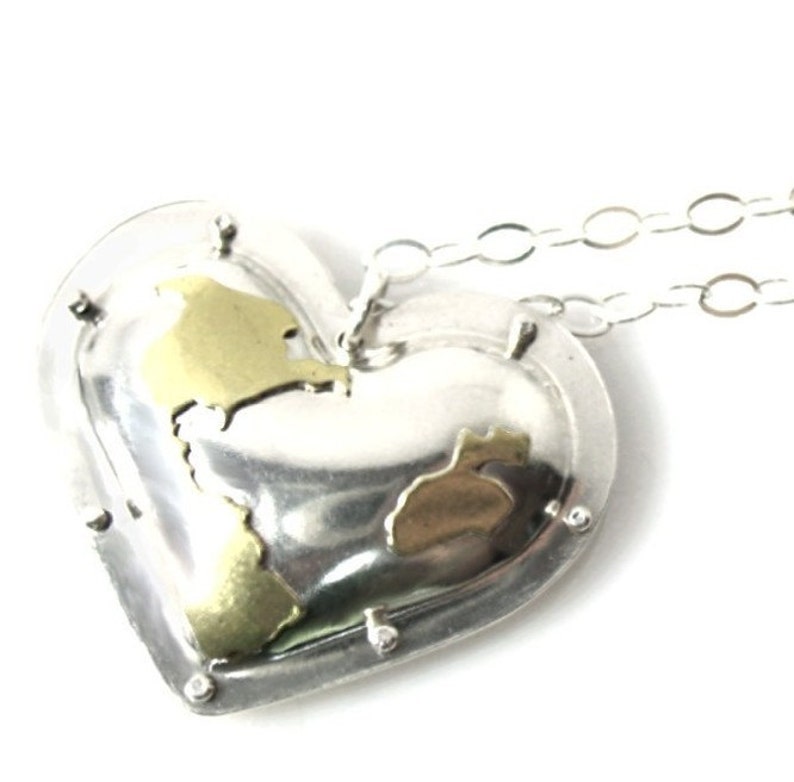 Puede incluir: Un collar con un colgante en forma de coraz&oacute;n plateado con un dise&ntilde;o de mapa dorado. El collar tiene una cadena delicada.