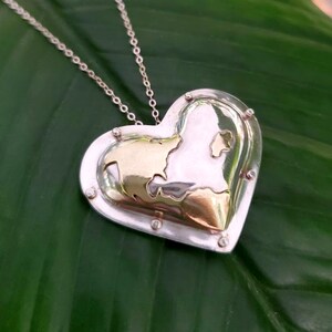 Puede incluir: Un collar con un colgante en forma de coraz&oacute;n de plata con un interior dorado. El colgante presenta un dise&ntilde;o texturizado y est&aacute; acentuado con peque&ntilde;os puntos plateados alrededor de los bordes.