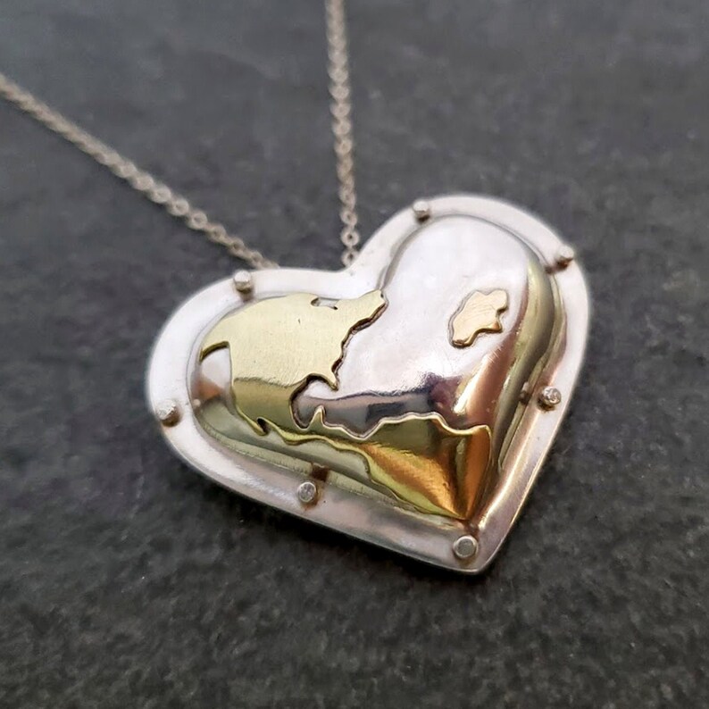 Puede incluir: Un collar con un colgante en forma de coraz&oacute;n plateado con un dise&ntilde;o de mapa dorado en el interior. El colgante tiene una superficie texturizada y est&aacute; acentuado con peque&ntilde;os tachuelas plateadas.
