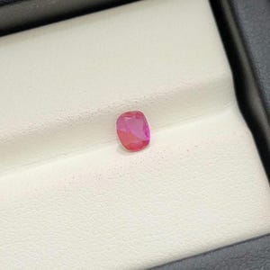 May include: A pink cushion-cut gemstone is displayed on a white surface inside a black box. The gem has a vivid colour and a polished surface. The contrasting setting enhances the gem's appearance, highlighting its brilliance and shape.