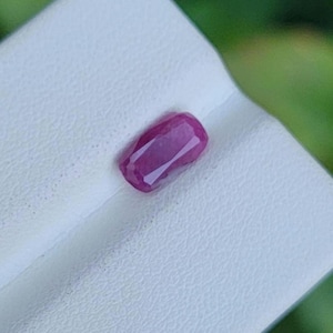 May include: A faceted, rectangular ruby gemstone with a deep pink hue. The gem is set on a white surface, with a blurred green background. The ruby's cut showcases its internal structure and light reflection.