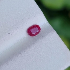 Peut inclure: Un rubis de forme ovale, d'un rouge profond. La gemme est posée sur une surface blanche, avec un arrière-plan vert flou. La surface polie du rubis reflète la lumière, soulignant sa clarté et sa couleur. Idéal pour la joaillerie.
