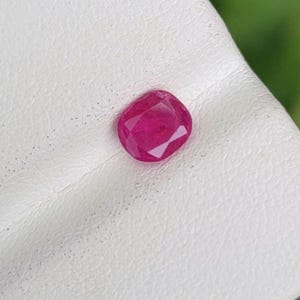 Pode incluir: Uma pedra preciosa de rubi facetada, em forma oval. O rubi é de uma cor vermelha profunda e vibrante e está colocado sobre uma superfície branca. O fundo é um verde desfocado.