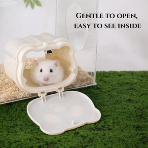 K&ouml;nnte beinhalten: Ein wei&szlig;er Hamster in einem beigefarbenen, b&auml;renf&ouml;rmigen Gehege mit offenem Deckel. Das Gehege befindet sich in einem transparenten Kunststoffhabitat mit Holzsp&auml;nen. Der Text lautet: &bdquo;GENTLE TO OPEN, EASY TO SEE INSIDE&ldquo;.