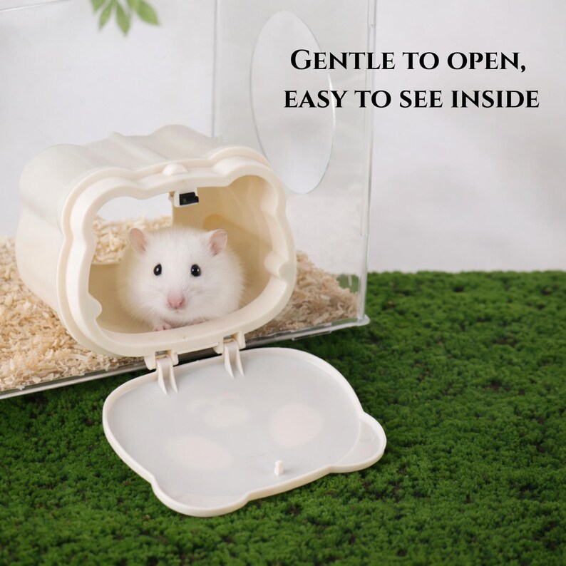 K&ouml;nnte beinhalten: Ein wei&szlig;er Hamster in einem beigefarbenen, b&auml;renf&ouml;rmigen Gehege mit offenem Deckel. Das Gehege befindet sich in einem transparenten Kunststoffhabitat mit Holzsp&auml;nen. Der Text lautet: &bdquo;GENTLE TO OPEN, EASY TO SEE INSIDE&ldquo;.