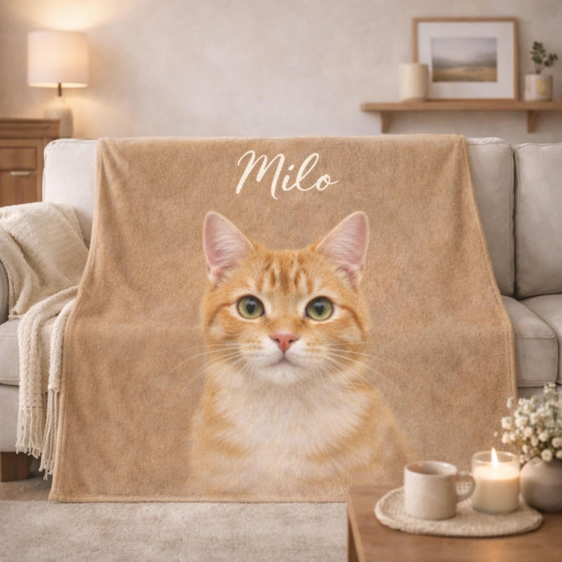 May include: A tan throw blanket featuring a large, detailed image of an orange tabby cat. The cat's name, "Milo," is written in cursive at the top. The blanket is draped over a light-colored sofa, creating a cozy and personalized home decor item.
