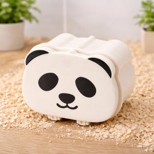 K&ouml;nnte beinhalten: Ein wei&szlig;er Beh&auml;lter in Pandaform mit schwarzen Augen, Ohren und Mund. Der Beh&auml;lter hat eine rechteckige Form mit abgerundeten Kanten und kleinen F&uuml;&szlig;en. Der Hintergrund ist eine hellbraune Oberfl&auml;che mit Holzsp&auml;nen.