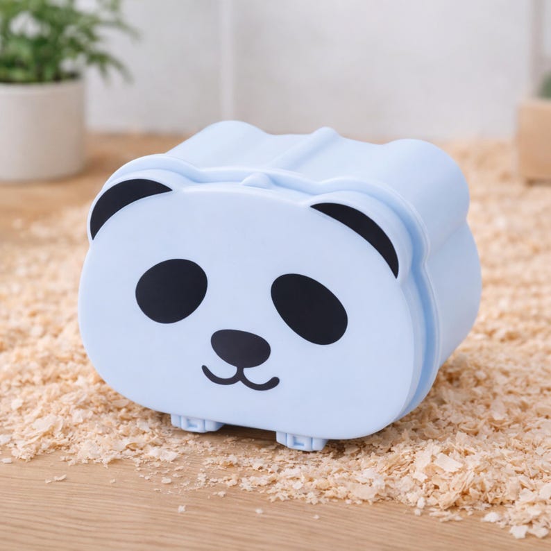 K&ouml;nnte beinhalten: Ein hellblaues Panda-f&ouml;rmiges Beh&auml;ltnis mit schwarzen Augen, Ohren und Nase. Der Beh&auml;lter hat eine rechteckige Form mit abgerundeten Kanten und kleinen F&uuml;&szlig;en. Der Hintergrund ist eine Holzoberfl&auml;che mit Holzsp&auml;nen.