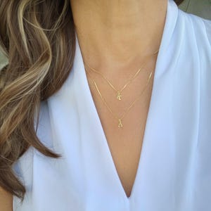 May include: Two delicate gold necklaces with initial pendants, one with the letter "A" and the other with the letter "K". The necklaces are layered and worn against a white blouse. The image is a close-up of the necklaces.