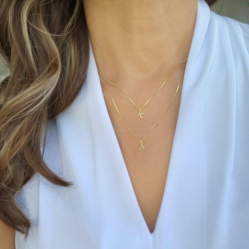 May include: Two delicate gold necklaces with initial pendants, one with the letter "A" and the other with the letter "K". The necklaces are layered and worn against a white blouse. The image is a close-up of the necklaces.