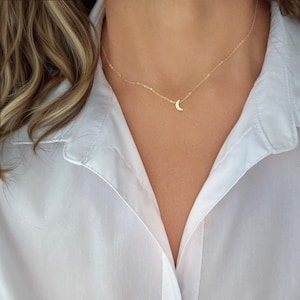 Puede incluir: Un delicado collar dorado con un colgante de media luna. El collar se lleva sobre una camisa blanca con cuello. La cadena es fina y el colgante de luna es pequeño y sencillo.