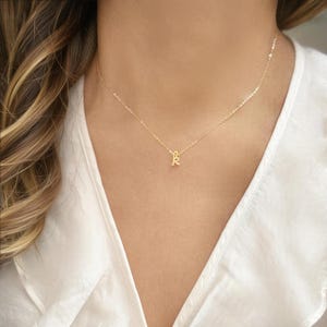 May include: A delicate gold-colored necklace featuring a small initial "K" pendant. The pendant hangs from a thin chain, resting on a person's chest. The background is a white blouse.