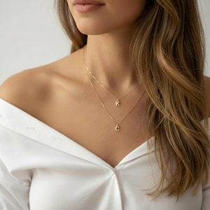 Könnte beinhalten: Zwei feine, goldene Halsketten, geschichtet, jeweils mit einem einzelnen Initialenanhänger. Der obere Anhänger ist ein "K", und der untere Anhänger ist ein "C". Die Halsketten werden von einer Person getragen, die ein weißes Hemd trägt.