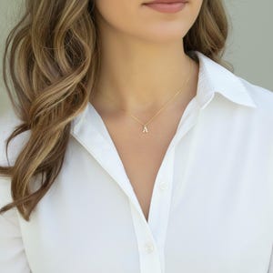 May include: A gold necklace featuring a small letter "A" pendant. The pendant is suspended from a fine chain. The necklace is worn over a white collared shirt.