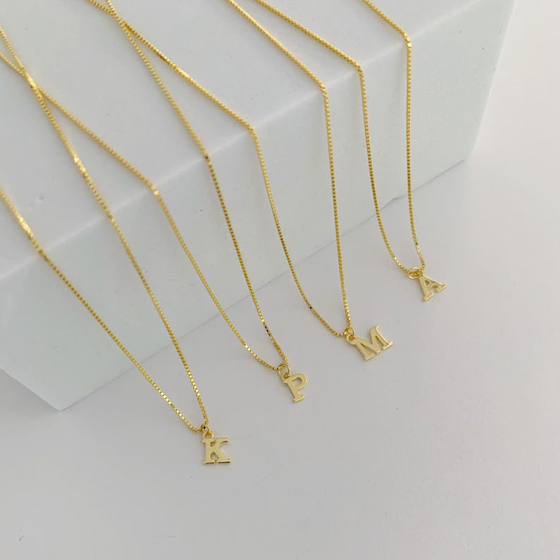 May include: Gold-coloured initial necklaces. Each necklace has a delicate chain and a small, block-letter initial pendant. The letters visible are K, P, M, and A. The necklaces are displayed on a white surface.