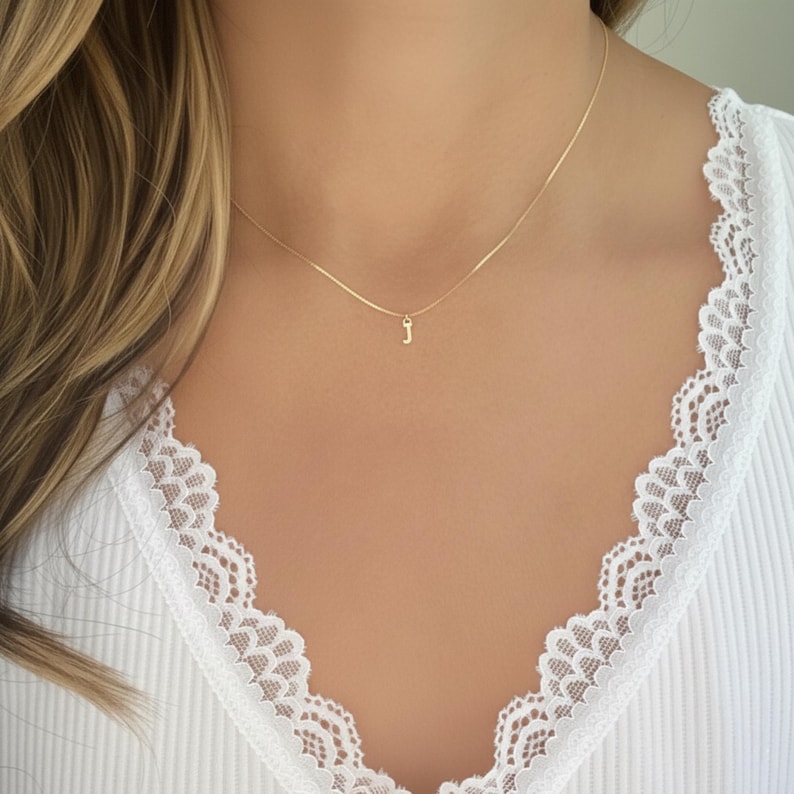 May include: A gold-coloured necklace with a small initial "J" pendant. The necklace is worn against a white ribbed top with lace trim. The chain is fine and the pendant is a simple, elegant design.