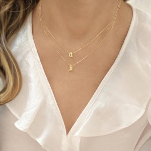 Könnte beinhalten: Zwei zarte, goldfarbene Halsketten, übereinander angeordnet, jeweils mit einem Anhänger mit einem einzelnen kursiven Buchstaben. Die obere Halskette zeigt den Buchstaben "a", die untere den Buchstaben "k". Die Halsketten werden vor einer weißen Bluse präsentiert.