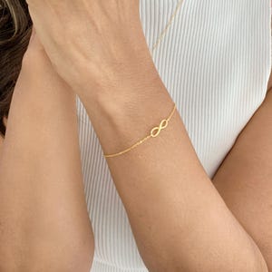 Könnte beinhalten: Ein goldenes Armband mit einem Unendlichkeits-Anhänger an einer zarten Kette. Das Armband wird am Handgelenk getragen.