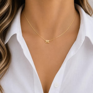 May include: A gold-toned necklace featuring a delicate chain and a single lowercase letter m pendant in a cursive script font. The necklace rests against a white collared shirt, showcasing a simple and refined design with a polished metallic finish.