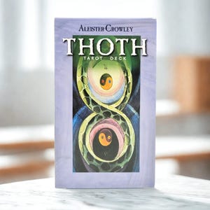 Op de afbeelding: De doos van het Aleister Crowley Thoth Tarot Deck heeft een lichtpaarse rand en een centrale illustratie van een groene slang die een oneindigheidssymbool vormt rond twee cirkelvormige yin-yang motieven in oranje, blauw en zwart.