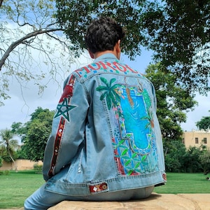 May include: Light blue denim jacket with colorful hand-painted artwork on the back and sleeves. The back features a tropical scene with palm trees and water. The sleeves have star and stripe designs. The word "MORO" is across the top of the back.