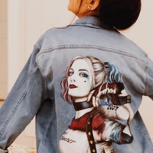 Può includere: Una giacca di jeans blu con un'immagine dipinta a mano di Harley Quinn sul retro. L'immagine è colorata e dettagliata, con il costume rosso e nero caratteristico di Harley Quinn, la sua mazza da baseball caratteristica e il suo trucco caratteristico. Il testo "Bad" e "Joker" è visibile sulla giacca.
