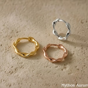 May include: Three wavy hoop earrings in gold, silver, and rose gold are displayed on a textured surface. The earrings have a unique, organic shape, and the colors create a striking contrast. The earrings are a stylish accessory.