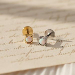 Puede incluir: Dos pendientes de botón con piedras cuadradas y opalescentes. Un pendiente es dorado, el otro plateado. Los pendientes se muestran sobre una superficie color crema con escritura.