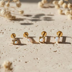 Può includere: Quattro orecchini a lobo dorati con pietre di opale bianche in varie forme: una stella, una marquise, un quadrato e una goccia. Gli orecchini sono esposti su una superficie chiara e strutturata, con fiori secchi sullo sfondo. Il marchio "Mythos Aurum" è in basso.