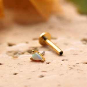 May include: A gold-coloured earring stud featuring a small, triangular opal stone and a gold post. The earring is on a textured, light brown surface. The stud has three small gold prongs securing the opal.