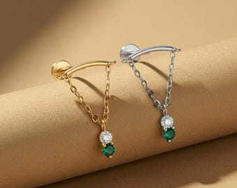 Peut inclure: Deux boucles d'oreilles, une dorée et une argentée, chacune avec une barre incurvée et une chaîne. Les chaînes suspendent une pierre transparente au-dessus d'une pierre verte. Les boucles d'oreilles sont présentées sur une surface beige. Le texte "Mythos Aurum" est en bas.