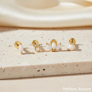 May include: Four gold-colored stud earrings with white opal stones. The earrings have different designs, including a teardrop shape, a cluster of small stones, and a floral design. The earrings are displayed on a textured, light-colored surface.