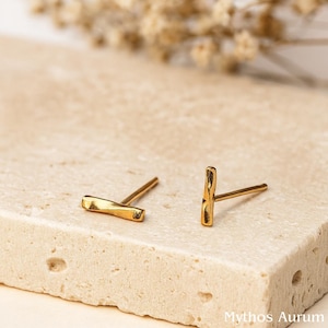 Peut inclure: Une paire de boucles d'oreilles à tige dorées. Les boucles d'oreilles ont un aspect texturé et martelé. Les boucles d'oreilles sont présentées sur une surface claire et texturée. La marque "Mythos Aurum" est visible en bas à droite.