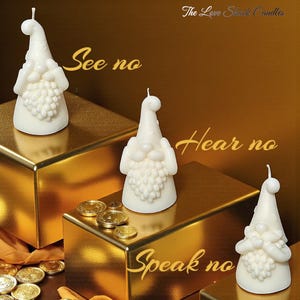 Può includere: Tre candele bianche a forma di gnomo, con barba e cappello strutturati, disposte su blocchi dorati. Le candele sono etichettate "See no", "Hear no" e "Speak no". Il logo The Love Shack Candles è in alto a destra.