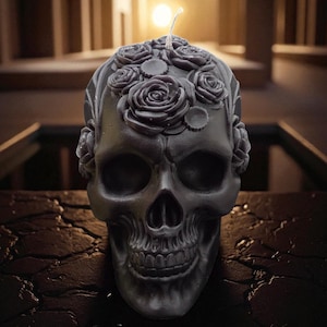 May include: A dark gray skull-shaped candle featuring a detailed arrangement of sculpted roses and small circular floral accents across the top and sides. A single white wick protrudes from the center of the floral crown on the skull's cranium.