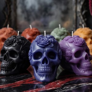 May include: A collection of skull-shaped candles featuring intricate molded rose details on the cranium. The candles are available in various solid colors including black, deep purple, light purple, orange, and green, each with a single white wick.
