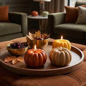 Puede incluir: Una bandeja redonda de madera contiene dos velas con forma de calabaza, una naranja y otra beige, con una vela de calabaza amarilla detrás. Un cuenco dorado contiene bayas oscuras y otro cuenco dorado contiene hojas de otoño secas.