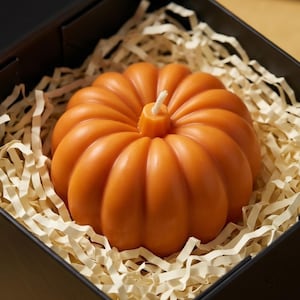 May include: A bright orange pumpkin-shaped candle with a central white wick, nestled inside a black box filled with light beige shredded paper filler. The candle features deep vertical ridges and a smooth, rounded surface texture.