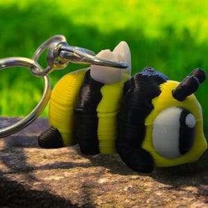 May include: A yellow and black bee-shaped keychain. The bee has a cartoonish design with a white eye and a silver keyring attached. The bee is resting on a stone surface with a blurred green background.