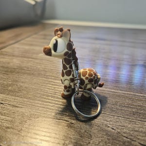 Op de afbeelding: Een giraf sleutelhanger met een bruin en wit gevlekt patroon. De giraf heeft grote ogen en is bevestigd aan een zilveren metalen sleutelring. De sleutelhanger is gemaakt van hard plastic.