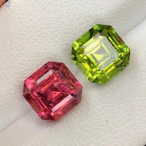 Può includere: Due gemme sfaccettate, una rosa-rossa vibrante e l'altra verde lime brillante, sono esposte su una superficie bianca. Le gemme, tagliate a forma quadrata con bordi smussati, catturano la luce e ne mostrano la chiarezza.