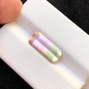 May include: A rectangular gemstone with a gradient of colors, including peach, pink, and green. The gemstone is set against a white textured surface. The gemstone is a natural crystal.