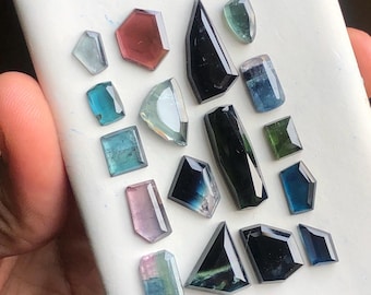 May include: A collection of various gemstone cuts in shades of blue, green, pink, and black. The gemstones are displayed on a white surface, showcasing their facets and clarity. The shapes include rectangles, triangles, and irregular forms.