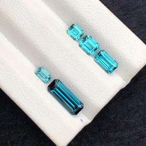 May include: Five loose, faceted gemstones in varying shades of teal and blue. The stones are cut in rectangular and square shapes, featuring emerald-style facets. They are arranged in two rows on a white, textured display surface with grooves.