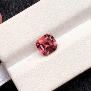 May include: A cushion-cut, pink gemstone rests on a white surface. The gemstone has facets that reflect light, creating a sparkling effect. The gem's color ranges from light to deep pink, with a hint of red.